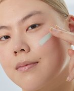 Close-up of a model with blonde hair swatching green color correcting makeup on her face