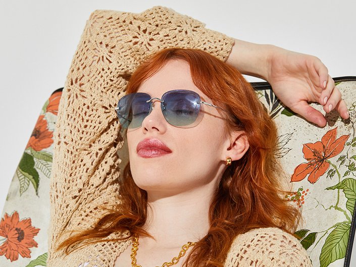 A person with red hair wearing blue sunglasses and a crocheted top lounges with their eyes closed on an orange floral beach chair