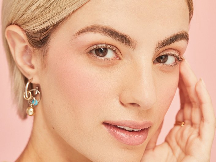 Person with a blonde pixie cut looks at the camera and touches their fingertips to their temple