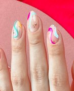 A close-up of a hand with nails painted in swirls of blue, purple, pink and yellow polish, accented with nail gems. 