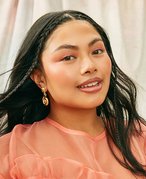person with long dark hair with two face-framing braids and peach-colored makeup matching a peach-colored shirt